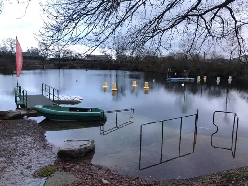 Plan d'eau calme avec une petite embarcation verte et des structures flottantes, arbres sans feuilles autour.
