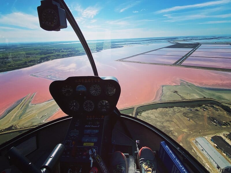 Vue depuis l'hélicoptère survolant des étendues d'eau colorées et des terres agricoles sous un ciel bleu clair.