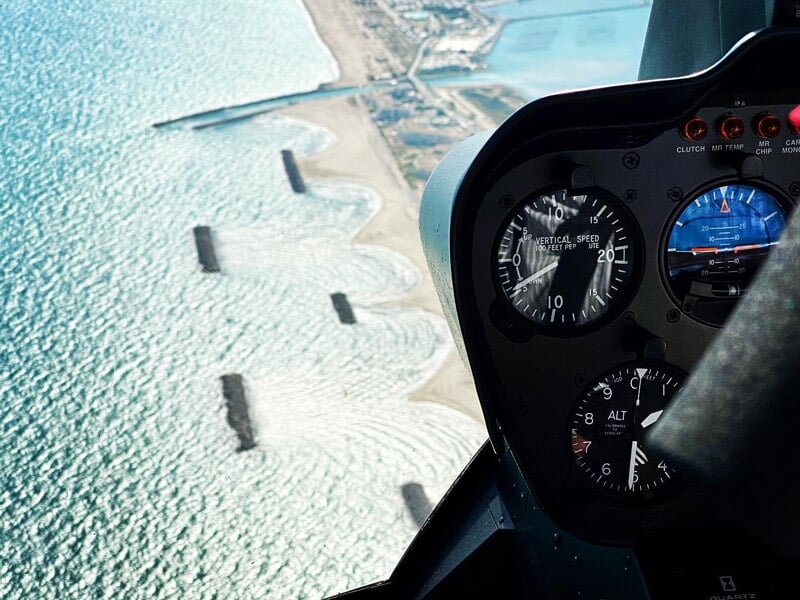 Cockpit d'hélicoptère avec instruments visibles, survolant une côte avec des structures en mer et une plage de sable.