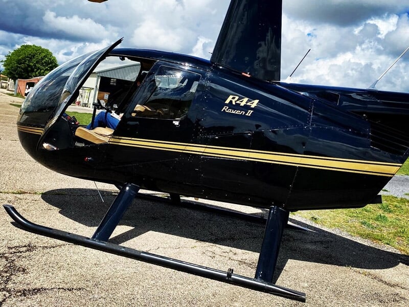 Hélicoptère noir R44 posé au sol sur une piste d'aérodrome, avec un ciel nuageux en arrière-plan.