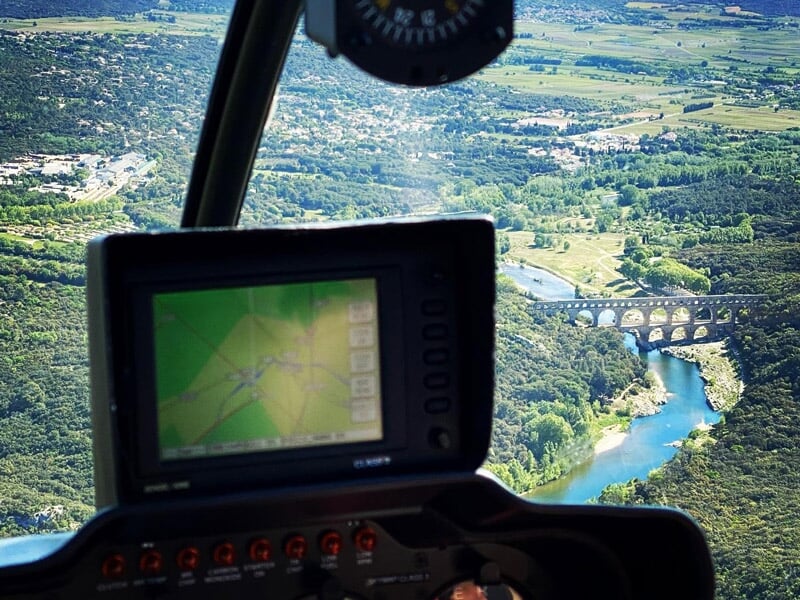 Vue depuis l'intérieur d'un hélicoptère avec écran GPS, survolant une rivière et un pont dans un paysage verdoyant.