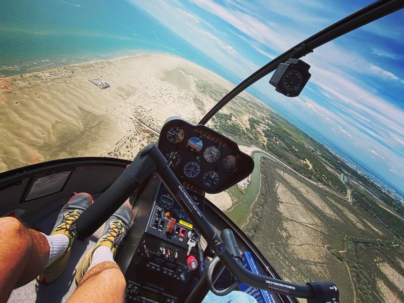 Vue depuis l'hélicoptère montrant les jambes du pilote et le tableau de bord, survolant un terrain désertique et vallonné.