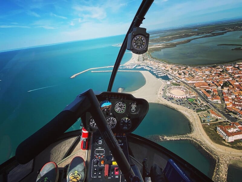 Cockpit d'hélicoptère avec instruments visibles, survolant une ville côtière avec plage, port et mer bleue.