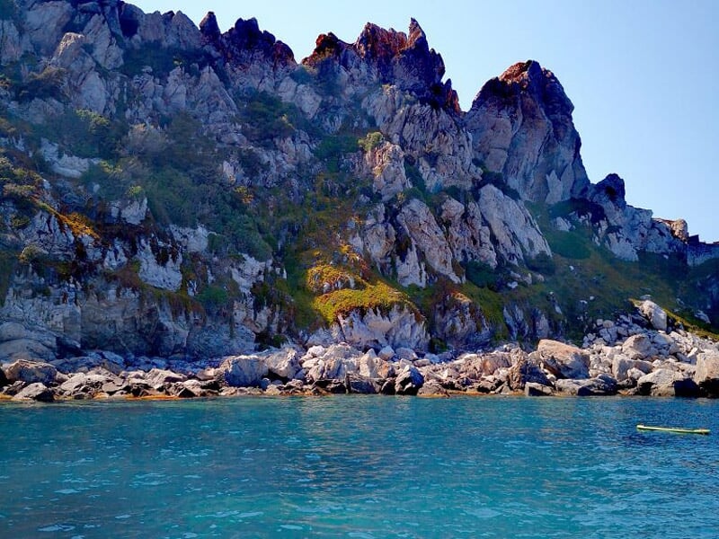 Vue d'une falaise rocheuse imposante au bord d'une mer calme et bleue sous un ciel clair ensoleillé.