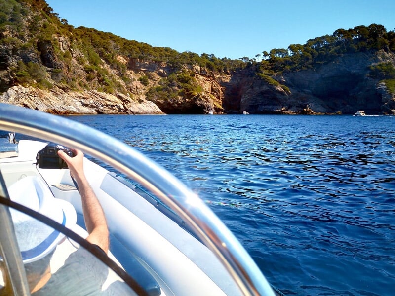 Personne à bord d'un bateau naviguant vers une crique rocheuse entourée d'arbres sous un ciel bleu.