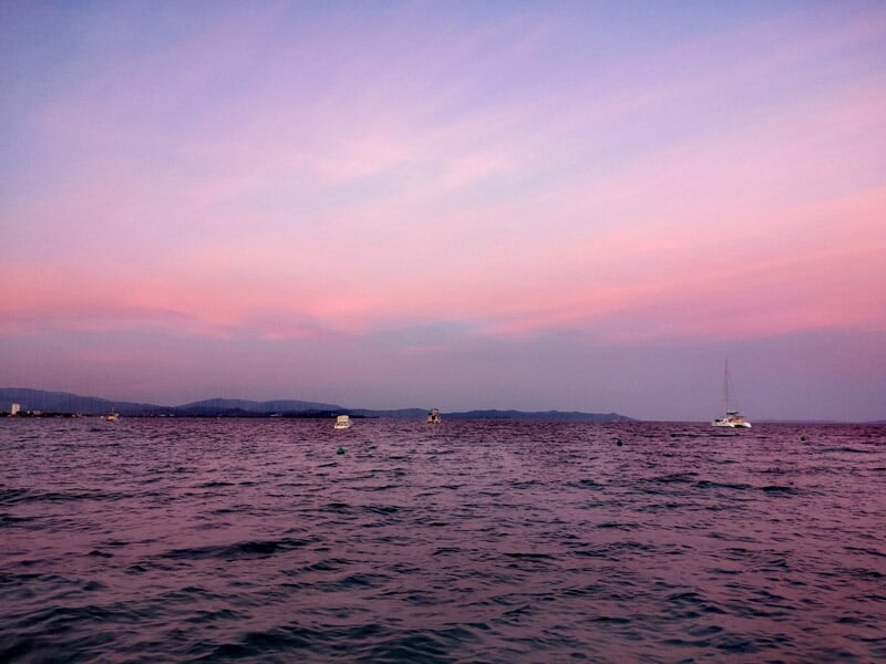 Coucher de soleil avec un ciel rose et violet au-dessus d'une mer calme où flottent plusieurs voiliers.