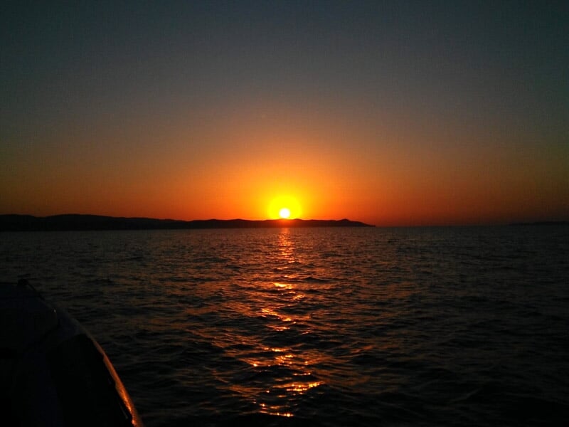 Soleil couchant rougeoyant à l'horizon sur une mer calme avec une silhouette de côte sombre en arrière-plan.
