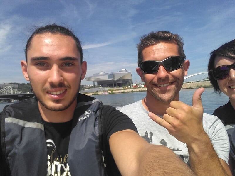 Trois personnes souriantes sur un bateau, avec un pont et un bâtiment en arrière-plan sous un ciel bleu.