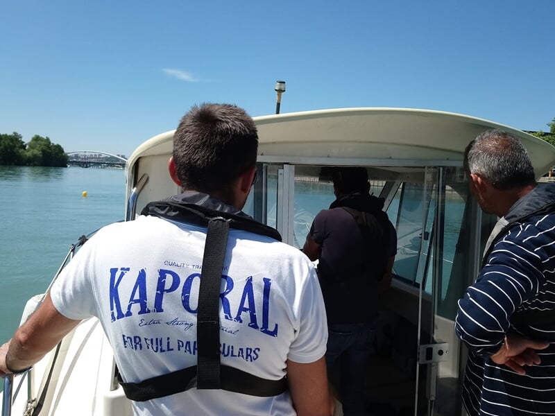 Trois hommes sur un bateau, l'un portant un t-shirt blanc avec un gilet de sauvetage, naviguant sur une rivière calme.