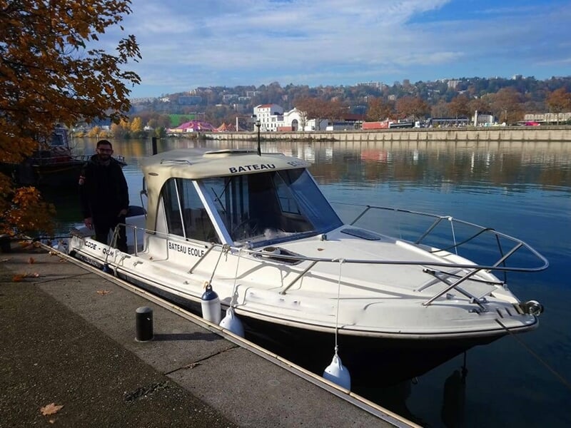 Bateau blanc amarré au bord d'un quai avec un homme debout à côté, paysage urbain en arrière-plan.
