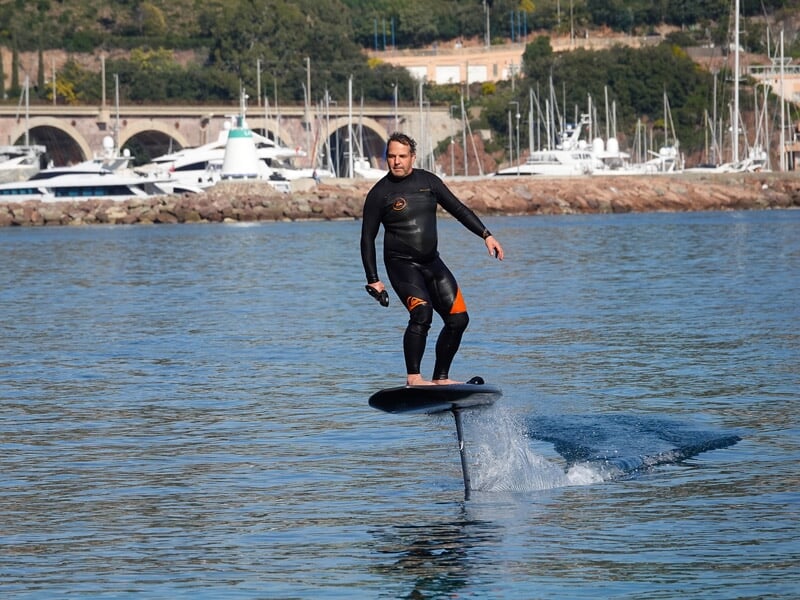 Homme en combinaison noire sur un surf électrique sur l'eau avec un port et des bateaux en arrière-plan.