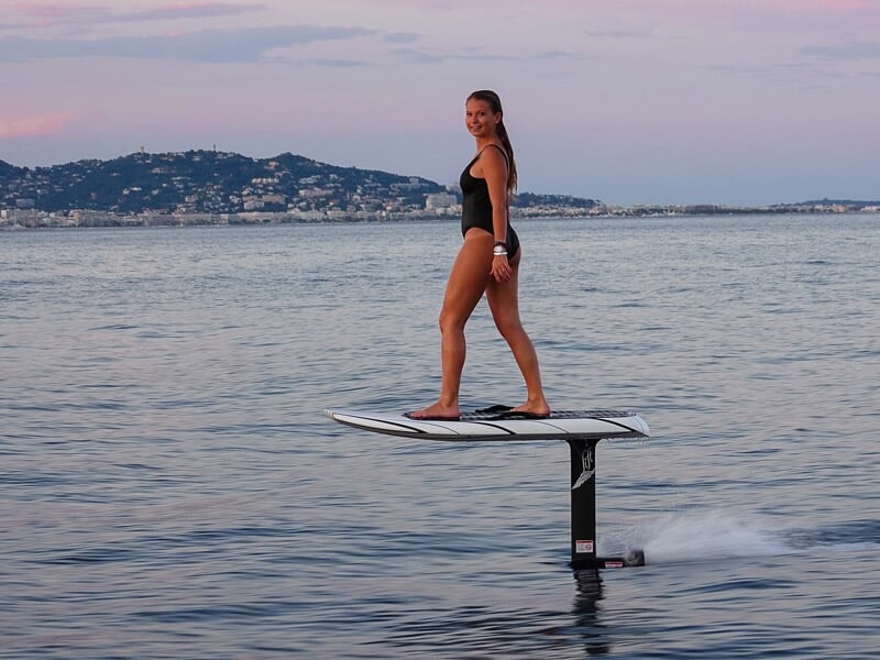 Femme en maillot noir debout sur un surf électrique sur une mer calme avec des collines en arrière-plan.