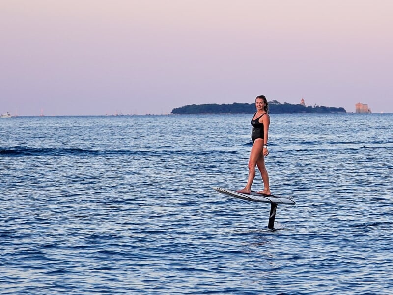 Femme souriante en maillot noir sur un surf électrique sur une mer calme au coucher du soleil.