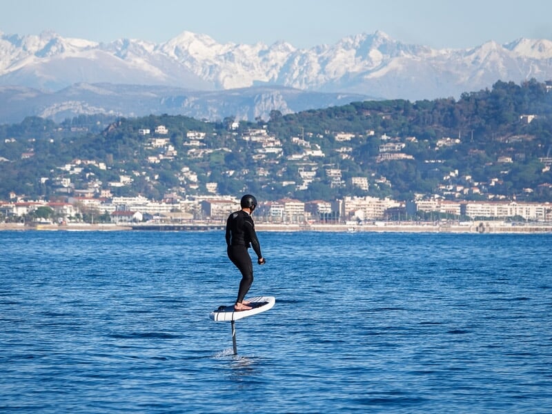 Homme en combinaison noire sur un surf électrique sur une mer calme avec une ville et montagnes enneigées en arrière-plan.