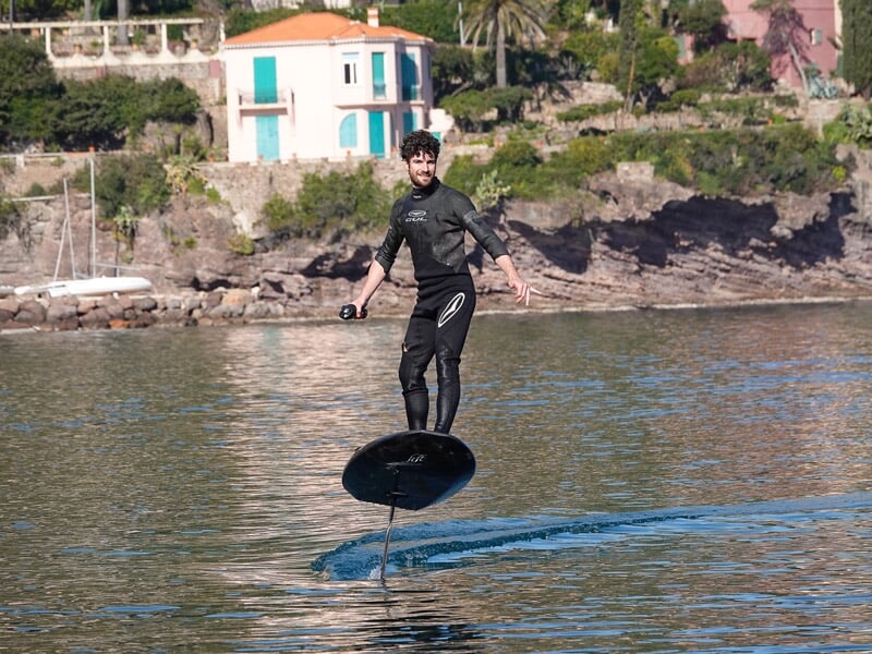 Homme en combinaison noire sur un surf électrique près d'une côte rocheuse avec une maison et des arbres.