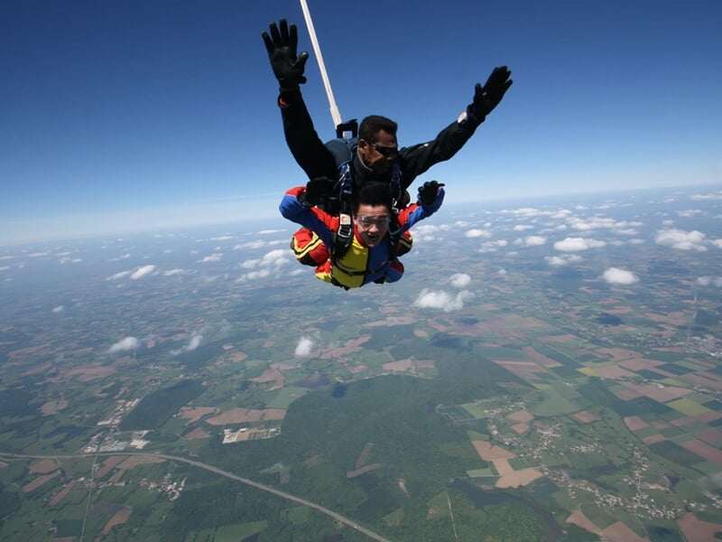 Deux parachutistes en tandem en chute libre au-dessus d'une campagne verte sous un ciel clair.