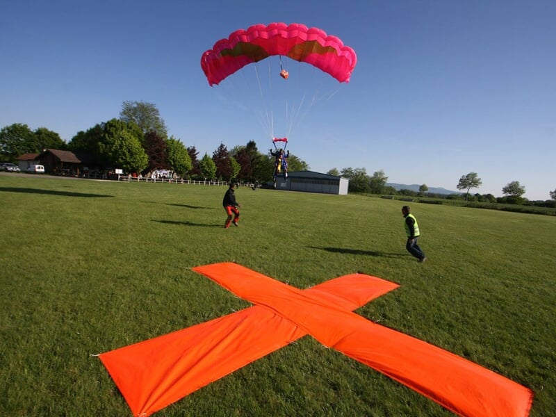 Parachutiste descendant avec un parachute rose au-dessus d'un champ vert avec des spectateurs au sol.