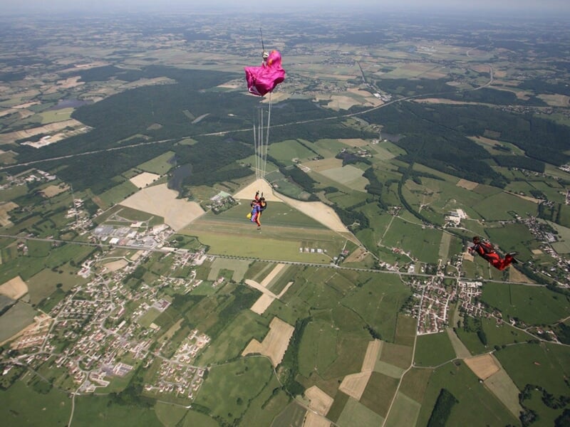 Vue aérienne d'un parachutiste avec parachute rose au-dessus d'un aérodrome entouré de champs et forêt.