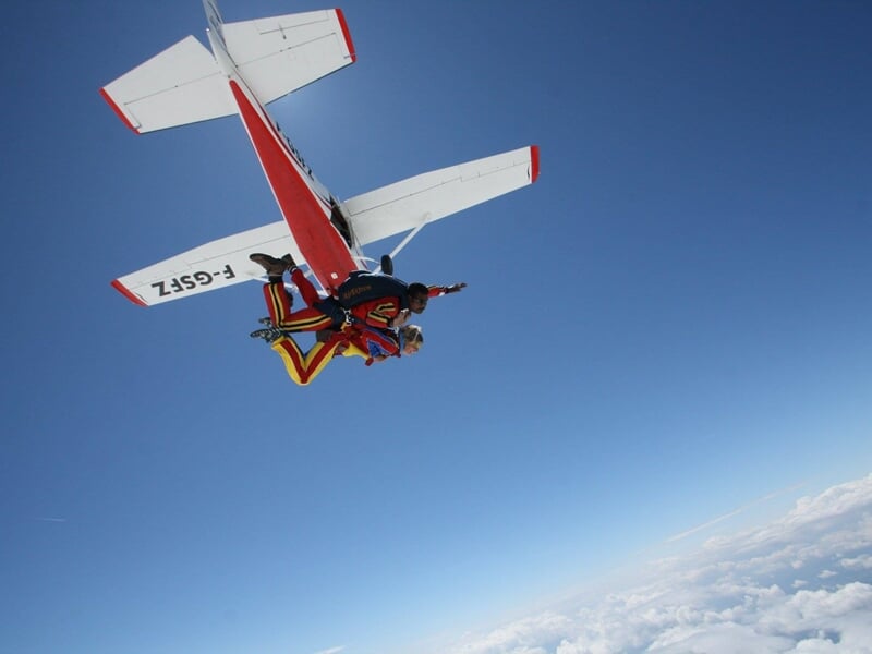 Saut en parachute en tandem depuis un avion blanc et rouge avec ciel bleu et nuages en arrière-plan.