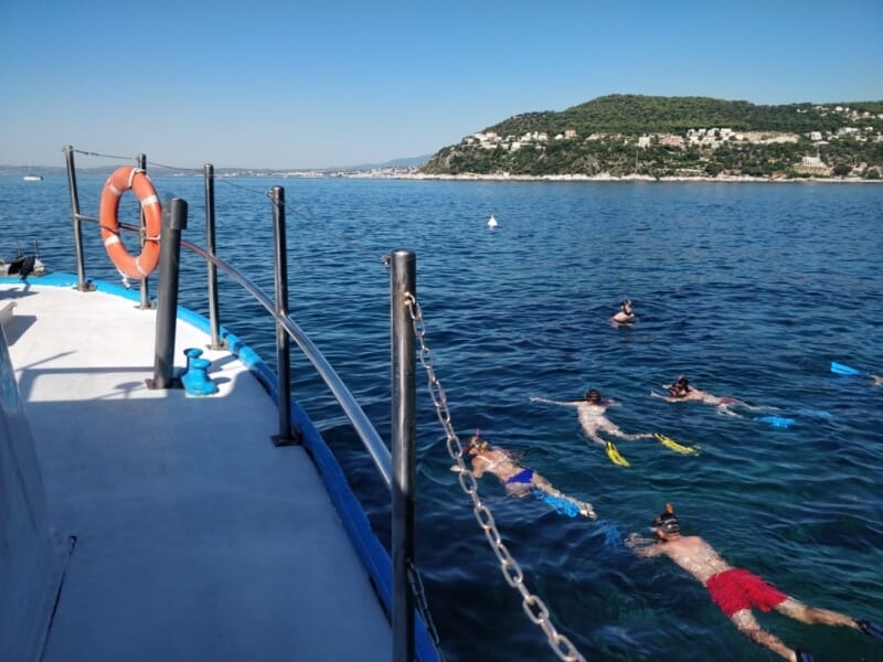 Groupe de personnes en snorkeling, équipées de palmes et de masques, nageant près d'un bateau en mer avec des collines verdoyantes en arrière-plan sous un ciel bleu dégagé.