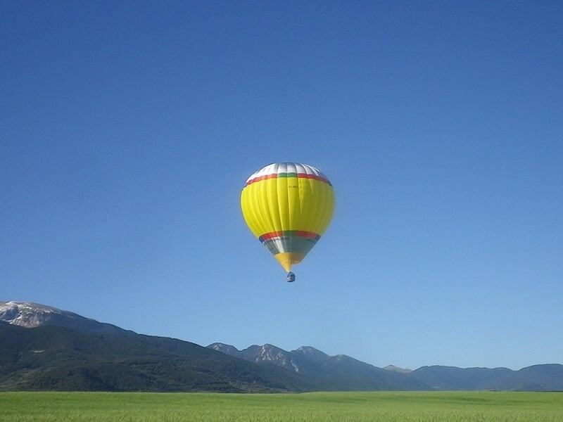 Montgolfière jaune avec une bande blanche et rouge survolant un champ vert avec des montagnes en arrière-plan.
