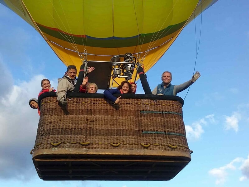 Groupe de six personnes souriantes dans la nacelle d'une montgolfière en vol sous un ciel partiellement nuageux.