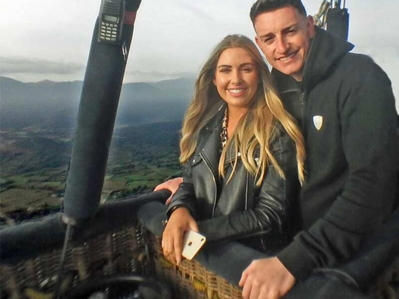 Jeune couple souriant dans la nacelle d'une montgolfière avec un paysage de collines et ciel nuageux en arrière-plan.