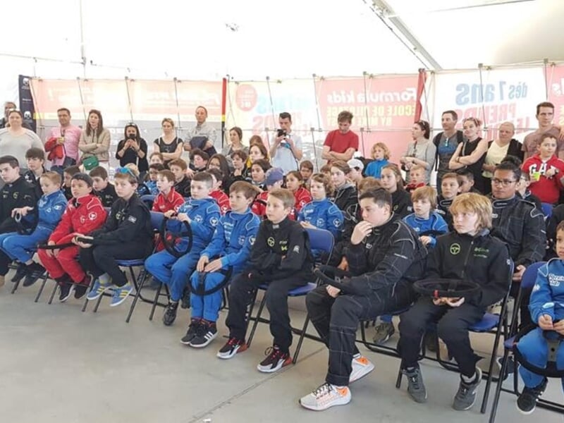 Enfants en combinaisons de course, assis et tenant des volants, entourés de parents et spectateurs dans une salle de formation.