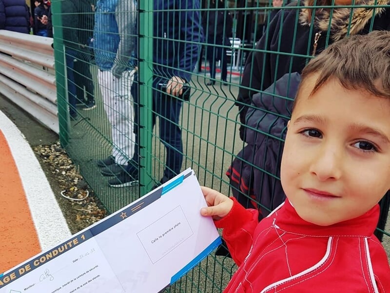 Petit garçon en combinaison rouge tenant un certificat de conduite près d'une piste de course.
