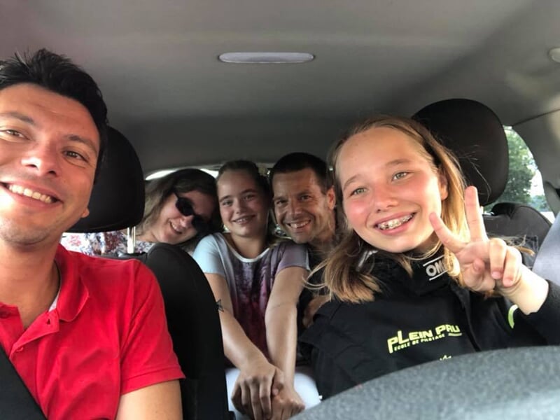 Groupe de cinq personnes souriantes dans une voiture, une fille faisant le signe de la paix au premier plan.