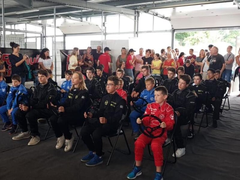 Enfants en combinaisons de course, assis et tenant des volants, entourés de parents et spectateurs dans une salle de formation.