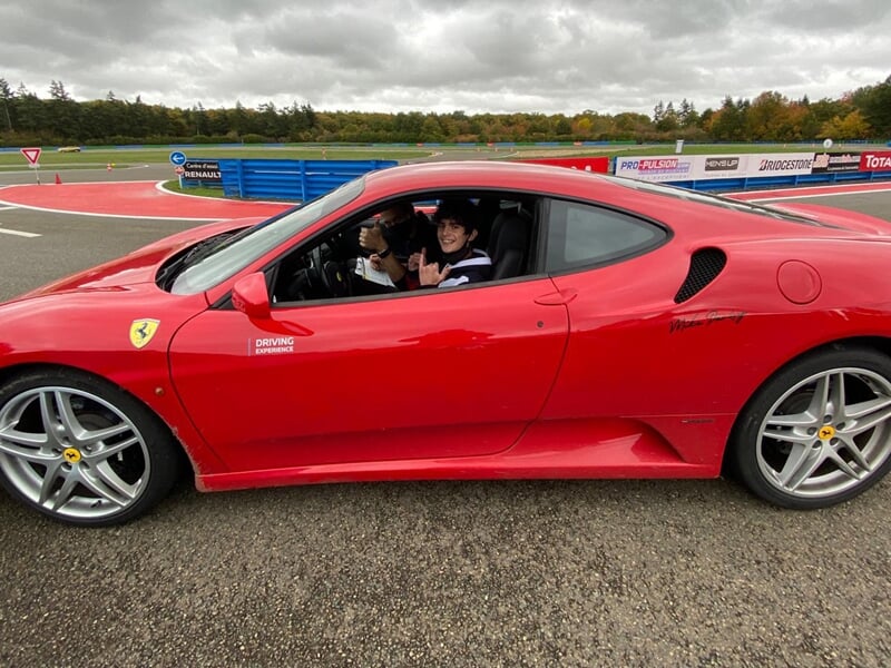Jeune homme souriant au volant d'une Ferrari rouge sur une piste de course, ciel nuageux.