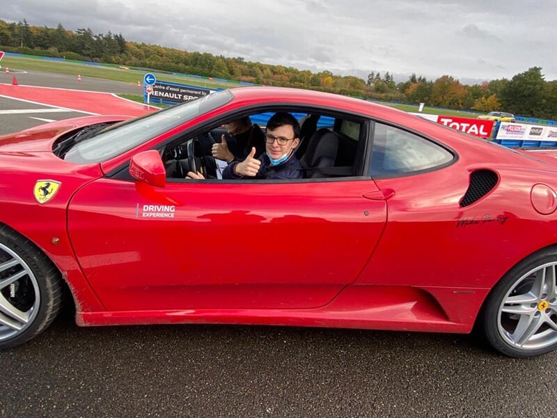 Adolescent au volant d'une Ferrari rouge, levant le pouce, sur une piste de course, ciel nuageux.