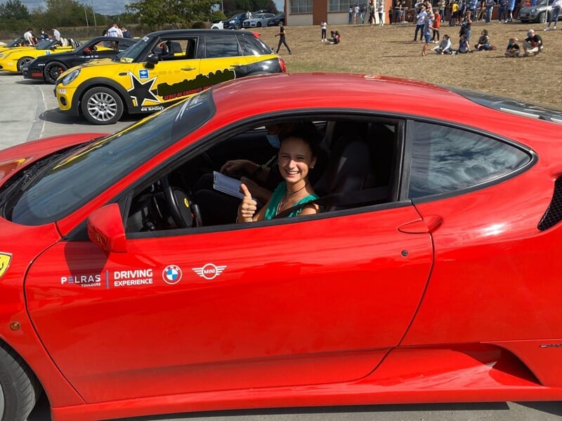 Femme souriante levant le pouce depuis l'intérieur d'une Ferrari rouge, Mini jaune en arrière-plan, sur une piste avec des spectateurs.