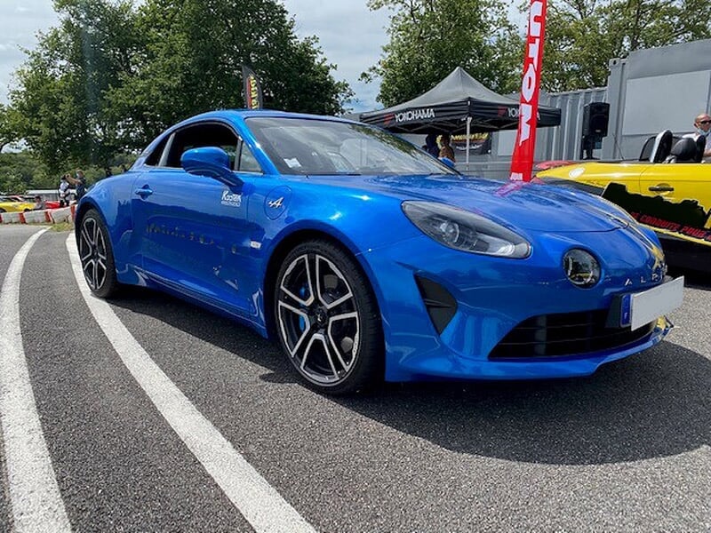 Alpine A110 bleue garée sur une piste de course, arbres et équipements de course en arrière-plan.