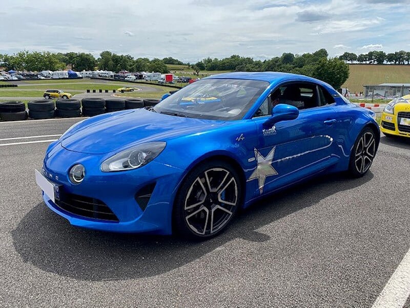 Alpine A110 bleue avec étoile argentée, vue de côté, garée sur une piste, pneus de sécurité et ciel nuageux en arrière-plan.