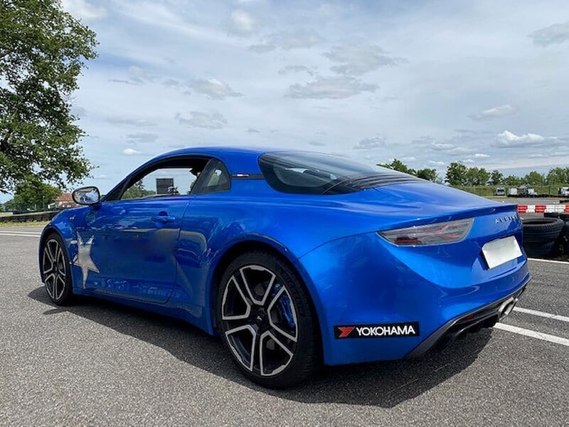 Alpine A110 bleue avec étoile argentée garée sur une piste, ciel partiellement nuageux en arrière-plan.