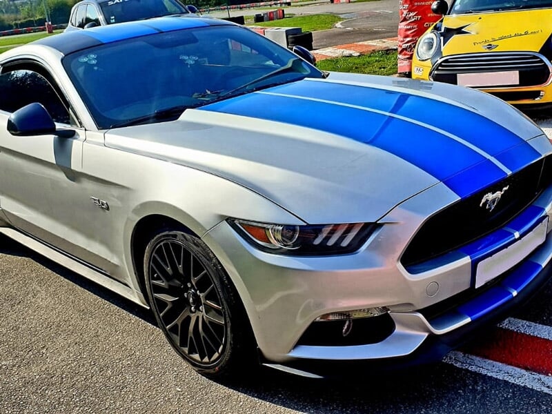 Ford Mustang V8 grise avec bandes bleues garée sur une piste de course, entourée d'autres véhicules colorés.