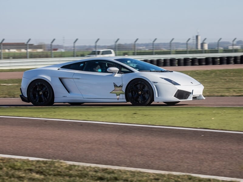 Lamborghini Gallardo LP560 blanche avec une étoile sur la portière, roulant sur une piste de course, clôture et paysage en arrière-plan.