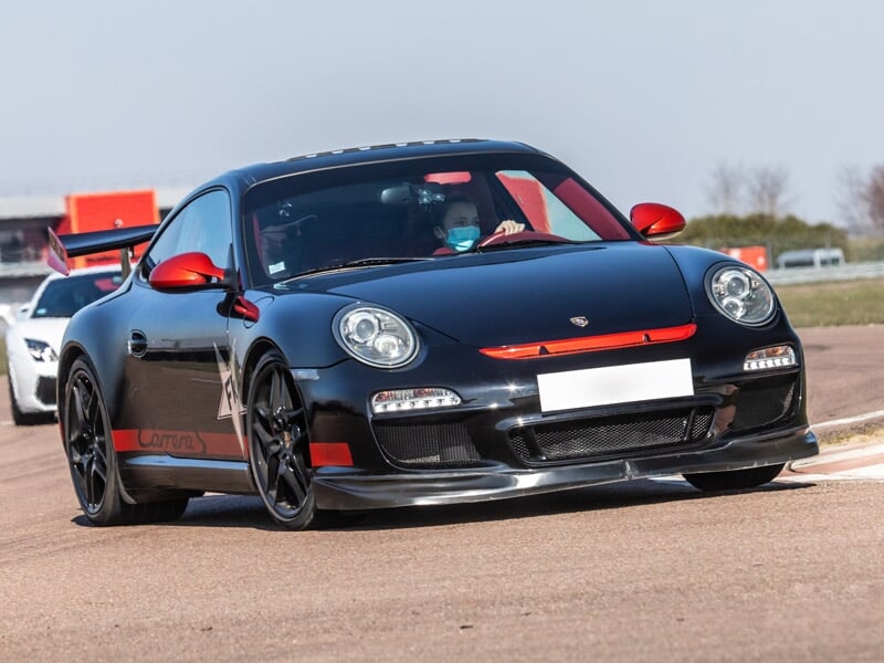 Porsche 997 Carrera S noire avec détails rouges sur une piste de course, deux personnes à l'intérieur, journée ensoleillée.