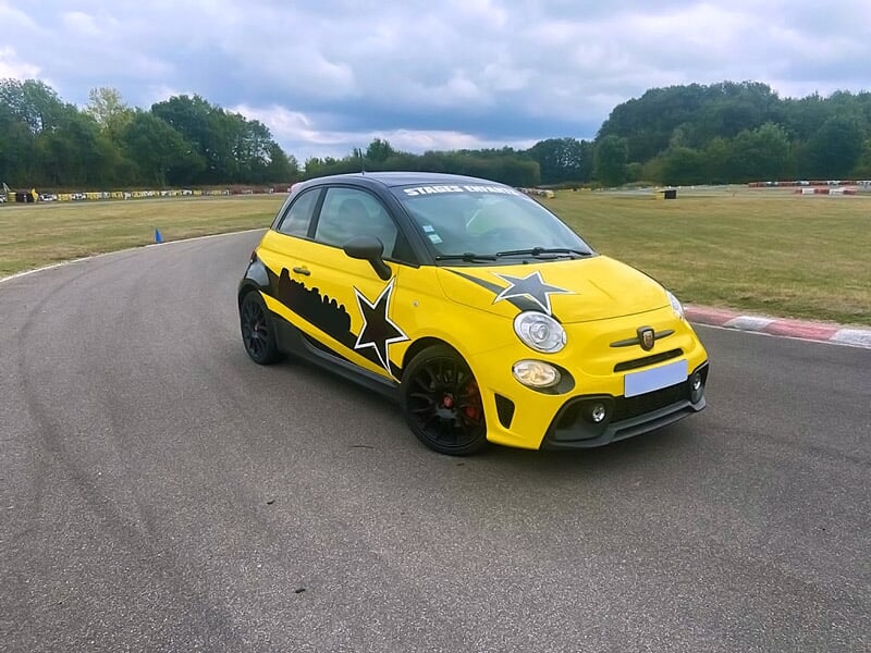 Fiat Abarth 500 Competizione jaune avec étoiles noires sur une piste de karting, végétation et ciel nuageux en arrière-plan.