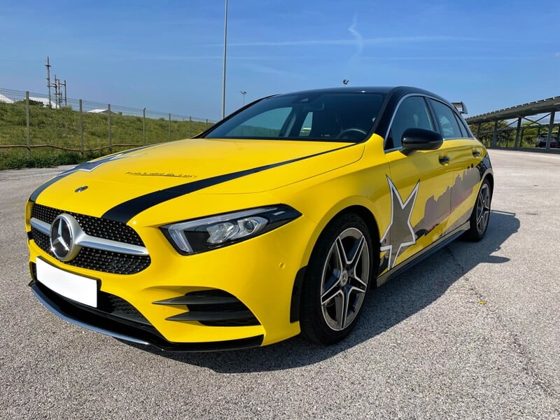 Mercedes Classe A AMG Line jaune avec étoiles grises et bandes noires garée sur une piste, ciel bleu clair en arrière-plan.