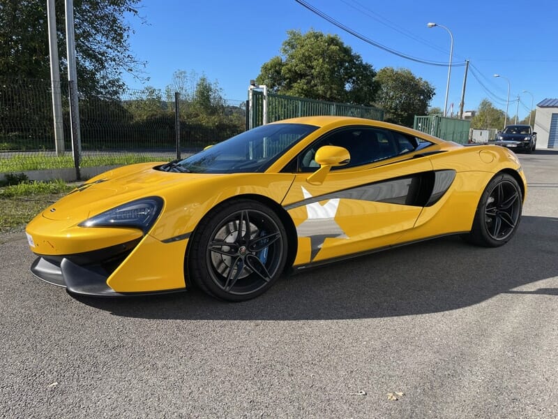 McLaren 540C Voiture de sport jaune et noire avec étoile grise garée sur une piste, arbres et clôtures en arrière-plan.