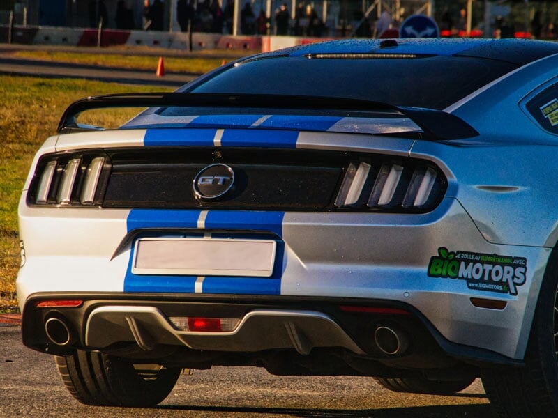 Ford Mustang GT blanche avec des bandes bleues. Publicité pour Big Motors sur le pare-chocs arrière. Circuit automobile avec barrières de sécurité en arrière-plan.