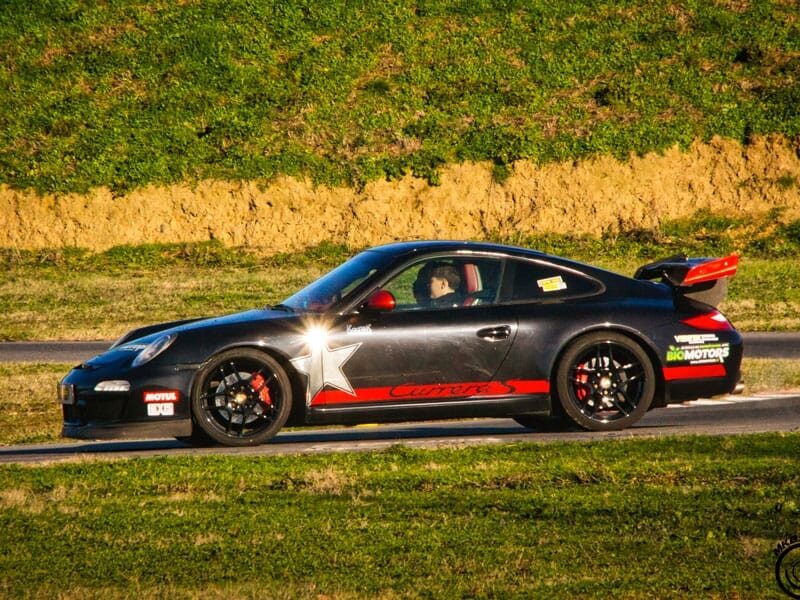 Porsche noire avec une étoile blanche sur la portière et des accents rouges. Publicités pour Motul et Big Motors. Circuit automobile avec herbe en arrière-plan.