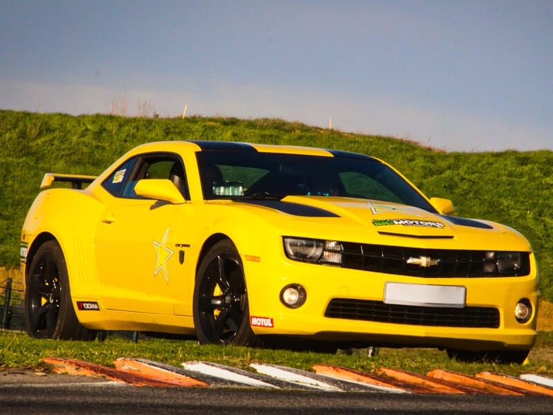 Chevrolet Camaro jaune avec des bandes noires et une étoile blanche sur la portière. Publicité pour Big Motors sur le capot. Circuit automobile avec bordure en arrière-plan.