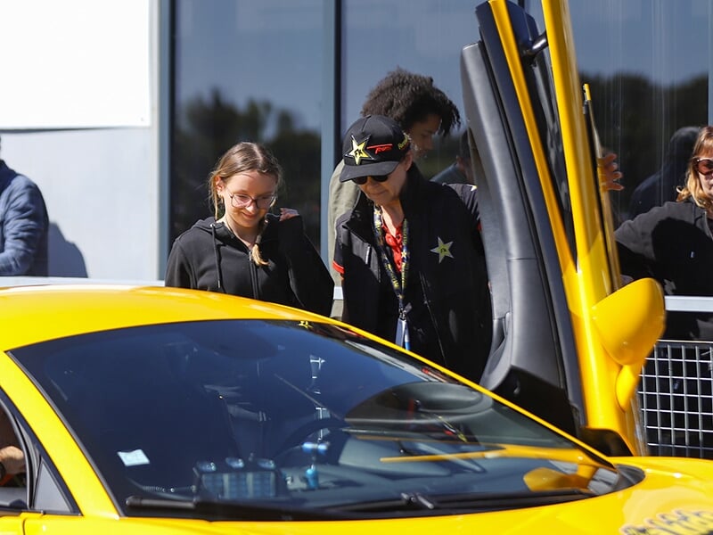 Une jeune fille souriante à côté d'une voiture jaune avec une porte papillon ouverte, accompagnée d'un instructeur en uniforme.