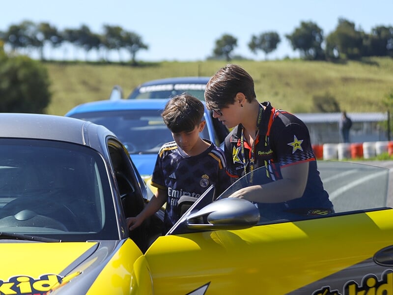 Enfant en maillot du Real Madrid écoutant une monitrice près d’une voiture sportive jaune sur un circuit. En arrière-plan, voitures de course et paysage verdoyant. Activité de pilotage ou stage de con