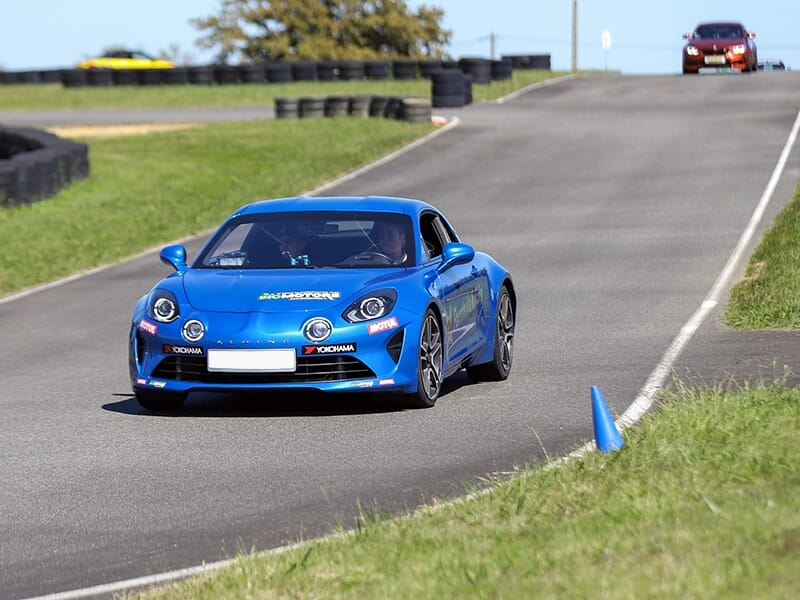 Une voiture bleue de sport en pleine vitesse sur un circuit de course, entourée de pneus de protection et de cônes de signalisation.