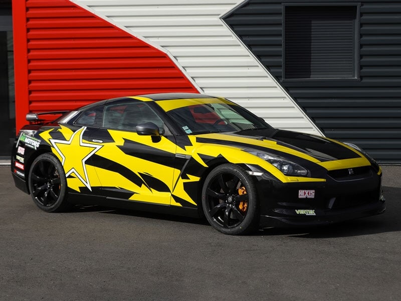 Une Nissan GTR jaune et noire est garée devant une porte de hangar rouge, blanche et noire